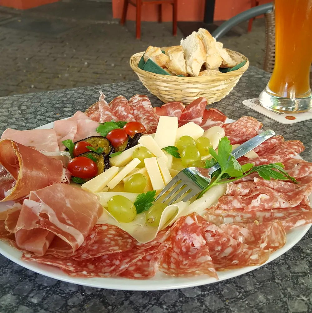 Teller mit Schinken, Salami, Käse, Tomaten und Weintrauben, dazu ein Brotkorb und ein Weizenbier